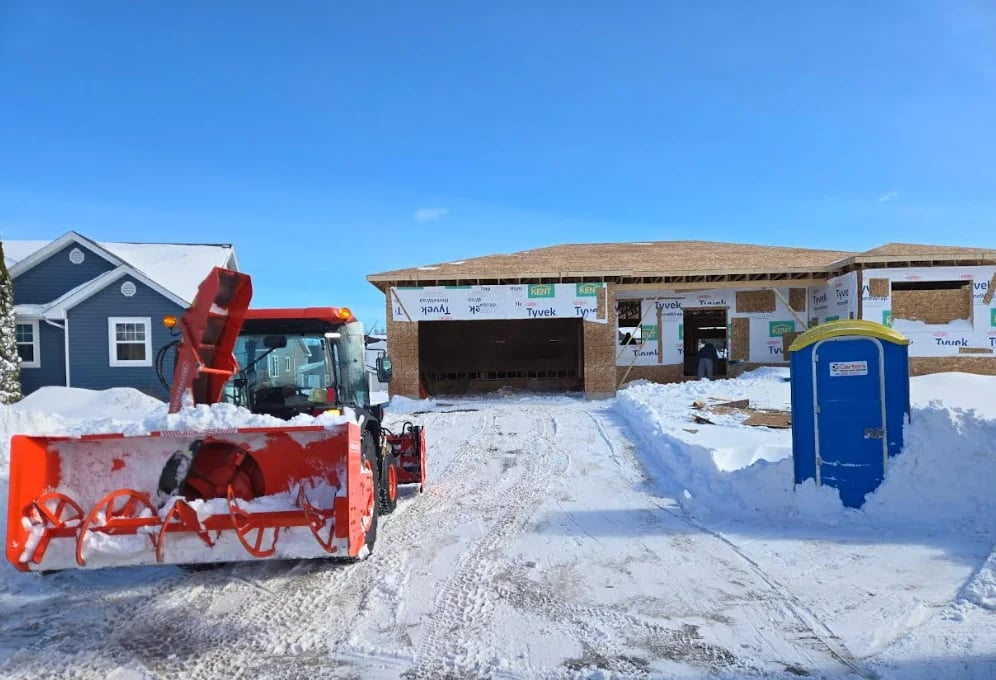 Red snow plow truck parked at snowy rural facility with buildings in background