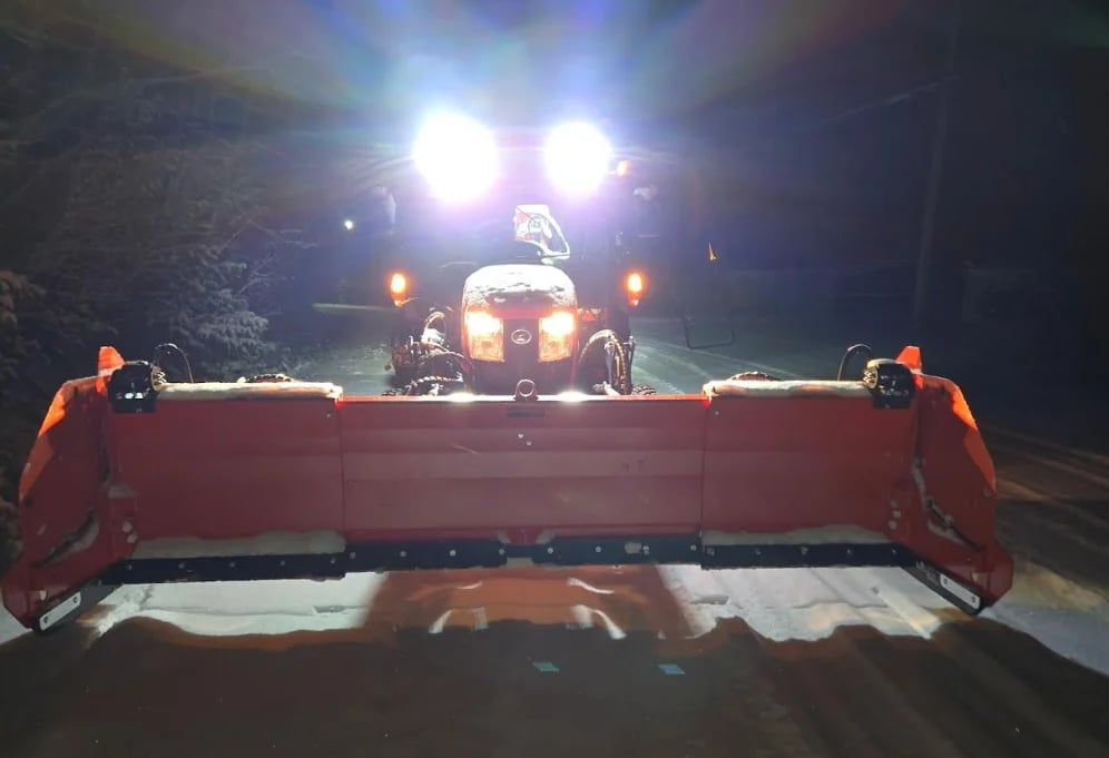 Red snow plow truck with bright headlights clearing a nighttime road, viewed from behind