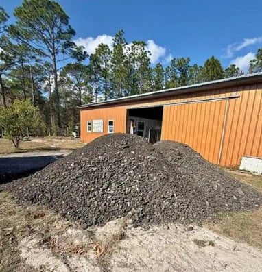 Large pile of crushed gravel in front of an orange and corrugated metal shed with tall pine trees in background
